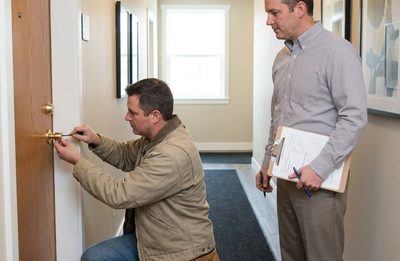 Locksmith replacing the lock cylinder on an apartment entry door while a property manager reviews paperwork